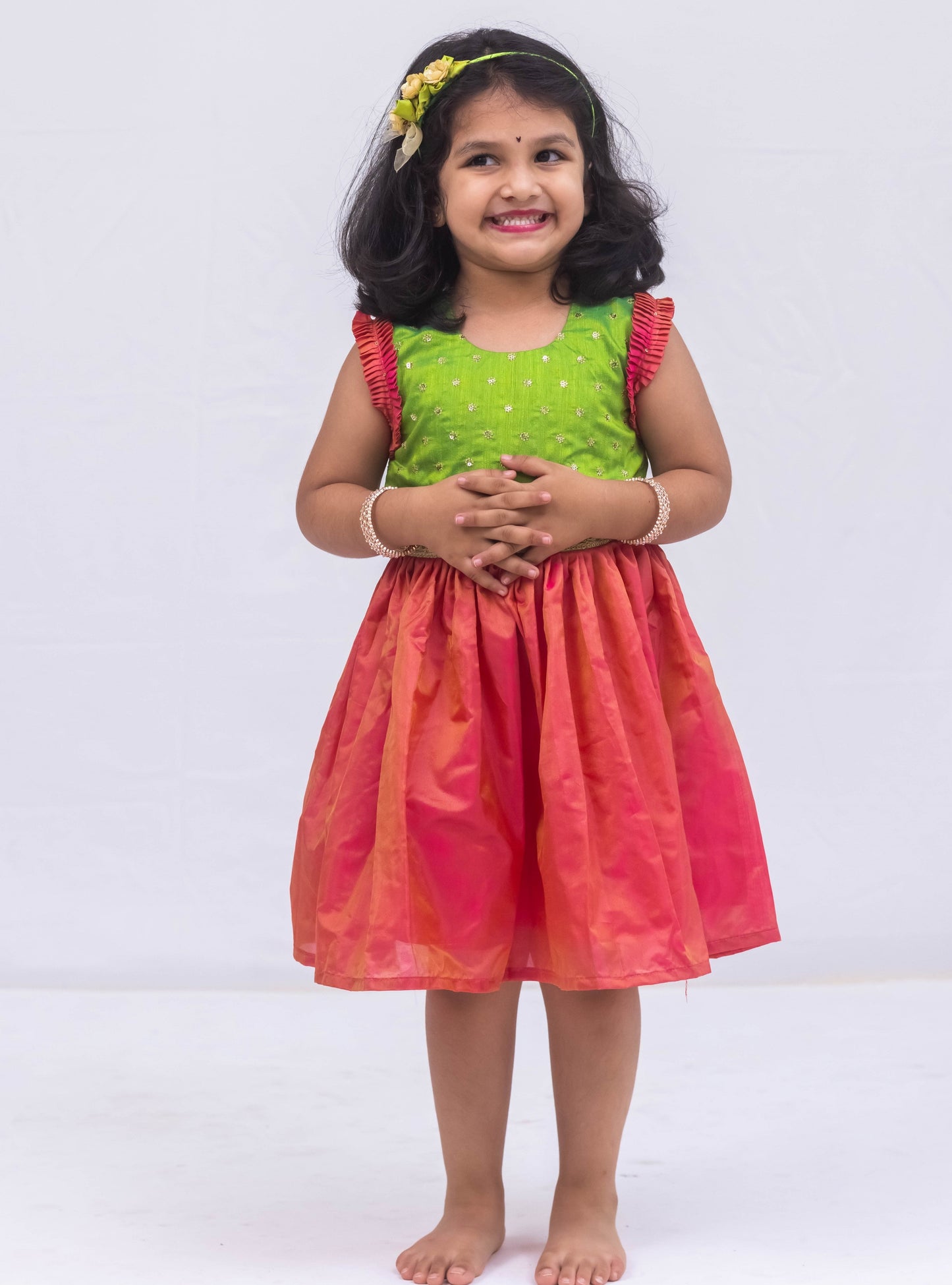 This exquisite frock for girls features a Parrot Green sequined bodice adorned with delicate lace detailing, paired with a luxurious Blush Pink Mysore silk skirt. The armholes are accentuated by intricately pleated frills that are adorned with matching sequins for a stunning finish.