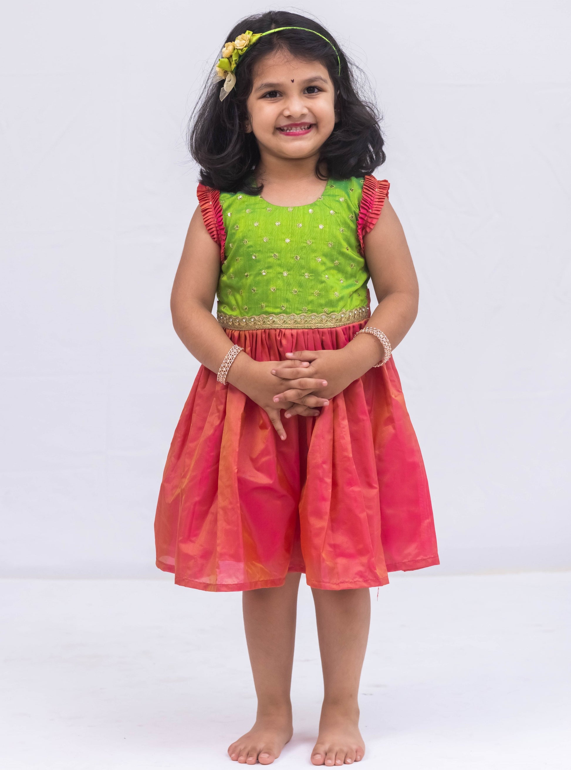 This exquisite frock for girls features a Parrot Green sequined bodice adorned with delicate lace detailing, paired with a luxurious Blush Pink Mysore silk skirt. The armholes are accentuated by intricately pleated frills that are adorned with matching sequins for a stunning finish.