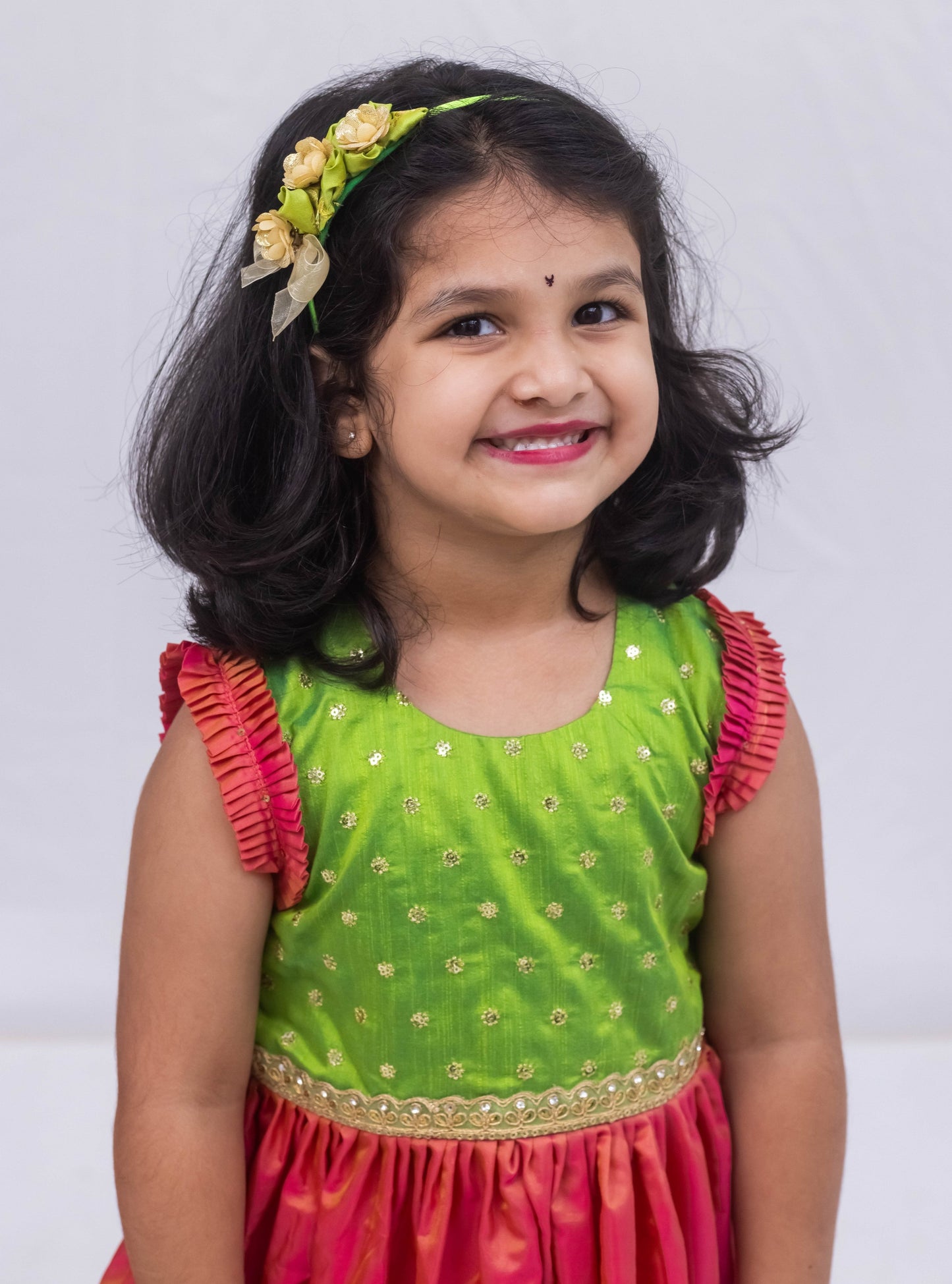 This exquisite frock for girls features a Parrot Green sequined bodice adorned with delicate lace detailing, paired with a luxurious Blush Pink Mysore silk skirt. The armholes are accentuated by intricately pleated frills that are adorned with matching sequins for a stunning finish.