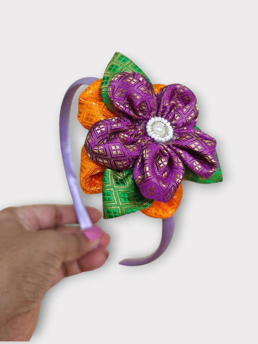 Magenta brocade layered flower motif , satin wrapped plastic hairband for Girls.This magenta brocade hairband features a layered flower motif and is wrapped in satin-covered plastic. It is perfect for girls aged two and up, making it a charming accessory for any outfit.